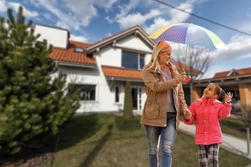 A woman and girl, holding hands, walk outside a house under a colorful umbrella. The girl wears a pink coat, and they both look up, smiling. Cloudy sky overhead.
